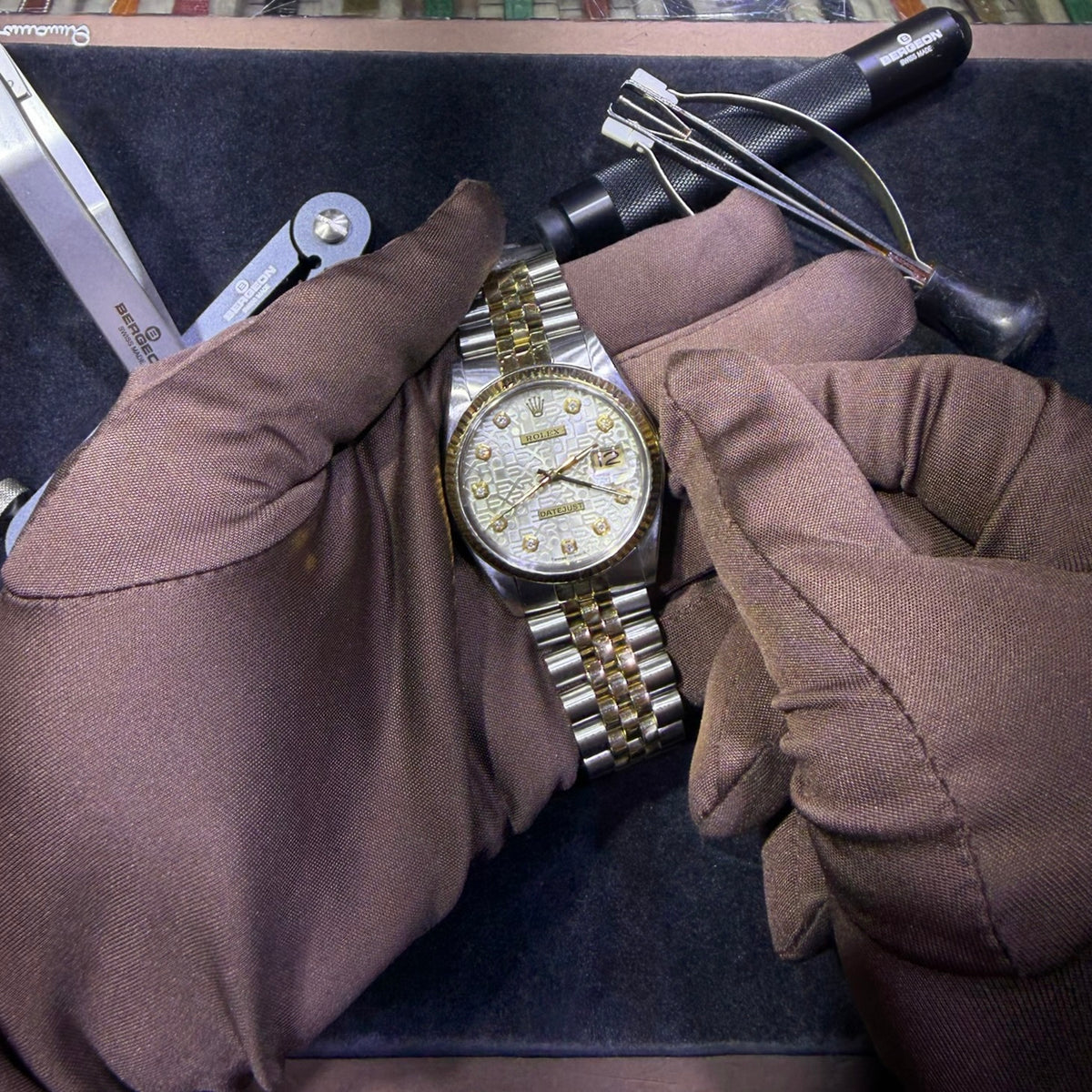 Gloved hands inspecting the dial and bracelet of a Rolex watch at the workbench.